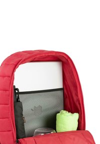 Mochila acolchada de color rojo con apertura de cremallera, que revela un bolsillo de malla y un objeto verde redondeado dentro, junto a un artículo negro no identificable.