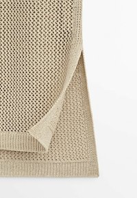 Tissu beige tricoté avec un motif à armure ouverte, plié pour montrer la texture et le bord rectangulaire ourlé sur un fond blanc.