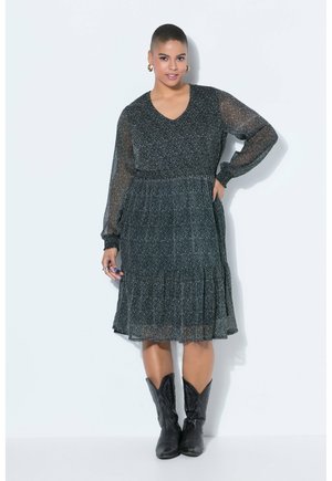 Donna con capelli corti che indossa un vestito nero a maniche lunghe con motivi, lungo fino al ginocchio, e stivali da cowboy neri, in piedi con una mano sui fianchi.