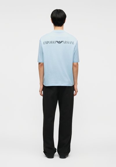 Άνδρας που φοράει ανοιχτό γαλάζιο κοντομάνικο πουκάμισο με το λογότυπο "EMPORIO ARMANI" στην πλάτη και μαύρο παντελόνι, στέκεται μπροστά σε μια απλή φόντο.