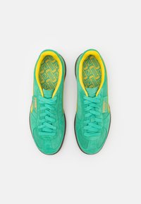 Une paire de baskets Puma teal avec une doublure intérieure jaune et un logo, des semelles intérieures à motifs et des semelles brunes, vues de dessus sur un fond blanc.