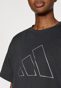 Μαύρο μπλουζάκι με στρογγυλό λαιμό και κοντά μανίκια, με μεγάλο ασημένιο λογότυπο Adidas σε γεωμετρικό σχέδιο στο μπροστινό μέρος.