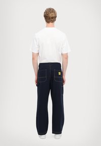 Navy cargo pants with wide legs, visible pockets, and contrasting orange stitching. Worn with a plain white t-shirt.
