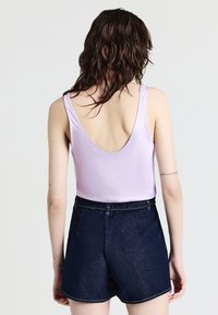 Donna con capelli castani ondulati che indossa una canotta lavanda a schiena bassa e shorts di jeans scuri, in piedi contro uno sfondo chiaro e uniforme.