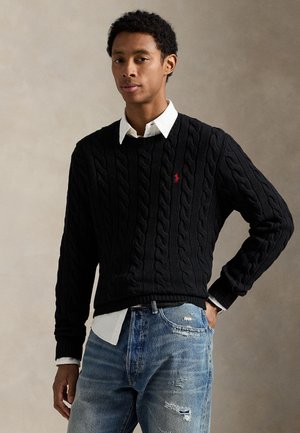 Schwarzer Pullover mit Zopfmuster, Rundhalsausschnitt und rotem Logo, kombiniert mit einem weißen Hemd mit Kragen und zerrissenen blauen Jeans.