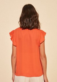 Blouse à manches courtes de couleur orange vif, avec des accents à volants et un tissu lisse. Vue de dos, avec une coupe droite et un ourlet arrondi.