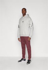 Sweatshirt cinza com capuz e três riscas pretas nas mangas, combinado com calças de treino bordeaux e sapatilhas pretas com detalhes brancos.
