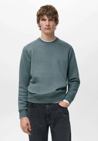 Giovane uomo dai capelli ricci castani che indossa una felpa blu petrolio con scollo rotondo e jeans neri, in piedi con una mano in tasca su sfondo bianco.