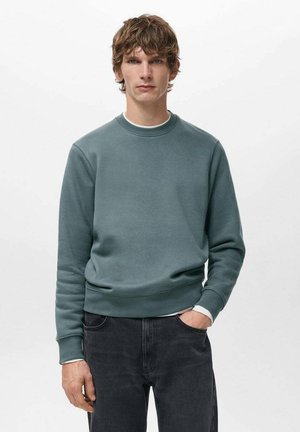 Giovane uomo dai capelli ricci castani che indossa una felpa blu petrolio con scollo rotondo e jeans neri, in piedi con una mano in tasca su sfondo bianco.