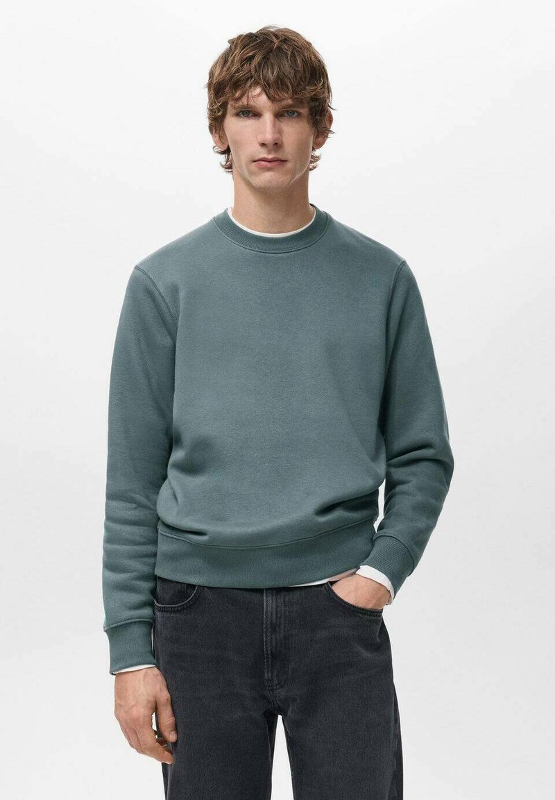 Giovane uomo dai capelli ricci castani che indossa una felpa blu petrolio con scollo rotondo e jeans neri, in piedi con una mano in tasca su sfondo bianco.