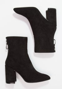 Bottines en suede noir avec un nez pointu, un talon bloc haut et un détail de fermeture éclair sur le côté. Texture lisse avec un minimum de coutures et sans branding visible.