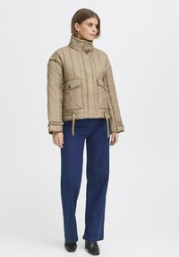 Beige quiltet jakke med høj krave, to forlommer og justerbare side remme, båret over mørkeblå bredbenede jeans.