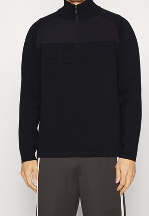 Mann trägt schwarzen Pullover mit Viertel-Reißverschluss und geprägtem "BOSS" auf der Brust sowie dunkle Hose mit weißen Seitenstreifen, steht vor einfarbigem Hintergrund.