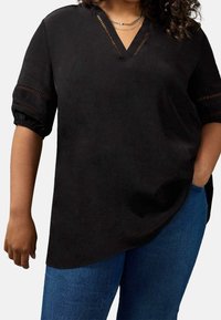 Donna che indossa una blusa nera a V con pizzo sulle maniche e sul collo, abbinata a jeans blu, con una mano in tasca su uno sfondo bianco.