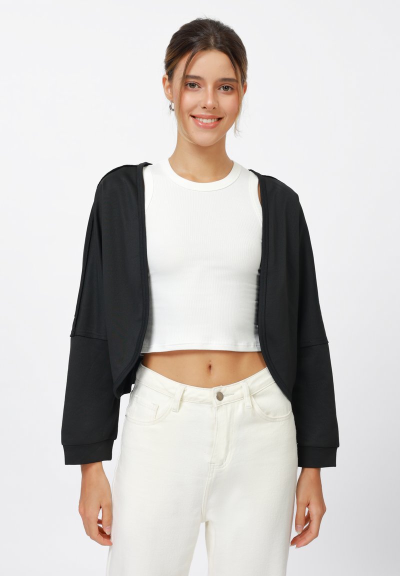 Veste noire courte avec une ouverture à l'avant, manches longues et texture lisse, portée sur un débardeur blanc ajusté et un pantalon taille haute blanc.