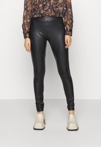 Legging en cuir synthétique noir, affichant un design ajusté, une texture lisse et des chevilles fuselées, associés à des chaussures à talons beiges.