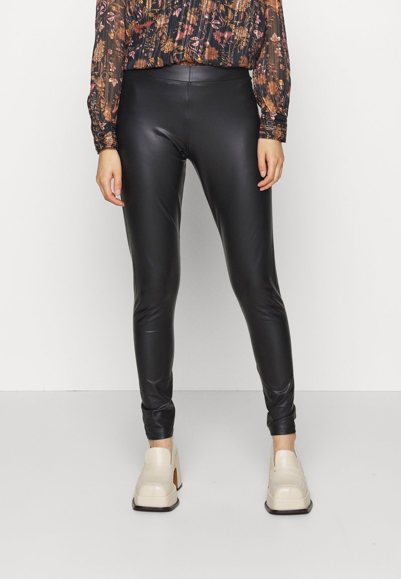 Legging en cuir synthétique noir, affichant un design ajusté, une texture lisse et des chevilles fuselées, associés à des chaussures à talons beiges.