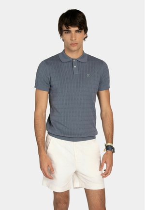 Młody mężczyzna ubrany w niebieską dzianinową koszulkę polo z krótkim rękawem i białe szorty, stojący na jednolitym tle.