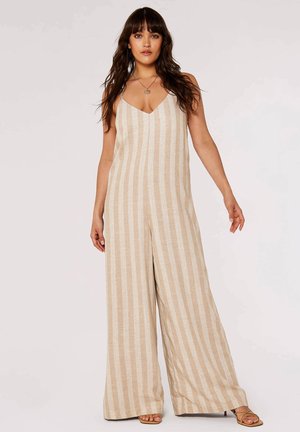 Frau in beiger und weißer, vertikal gestreifter, ärmelloser, weiter Bein-Jumpsuit mit V-Ausschnitt und Sandalen, stehend vor einem schlichten Hintergrund.