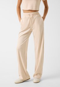 Beige Jersey-Hose mit einem Kordelzugbund, Seitentaschen und einem leichten Glanz, kombiniert mit weißen Sneakers. Elegante und entspannte Passform.
