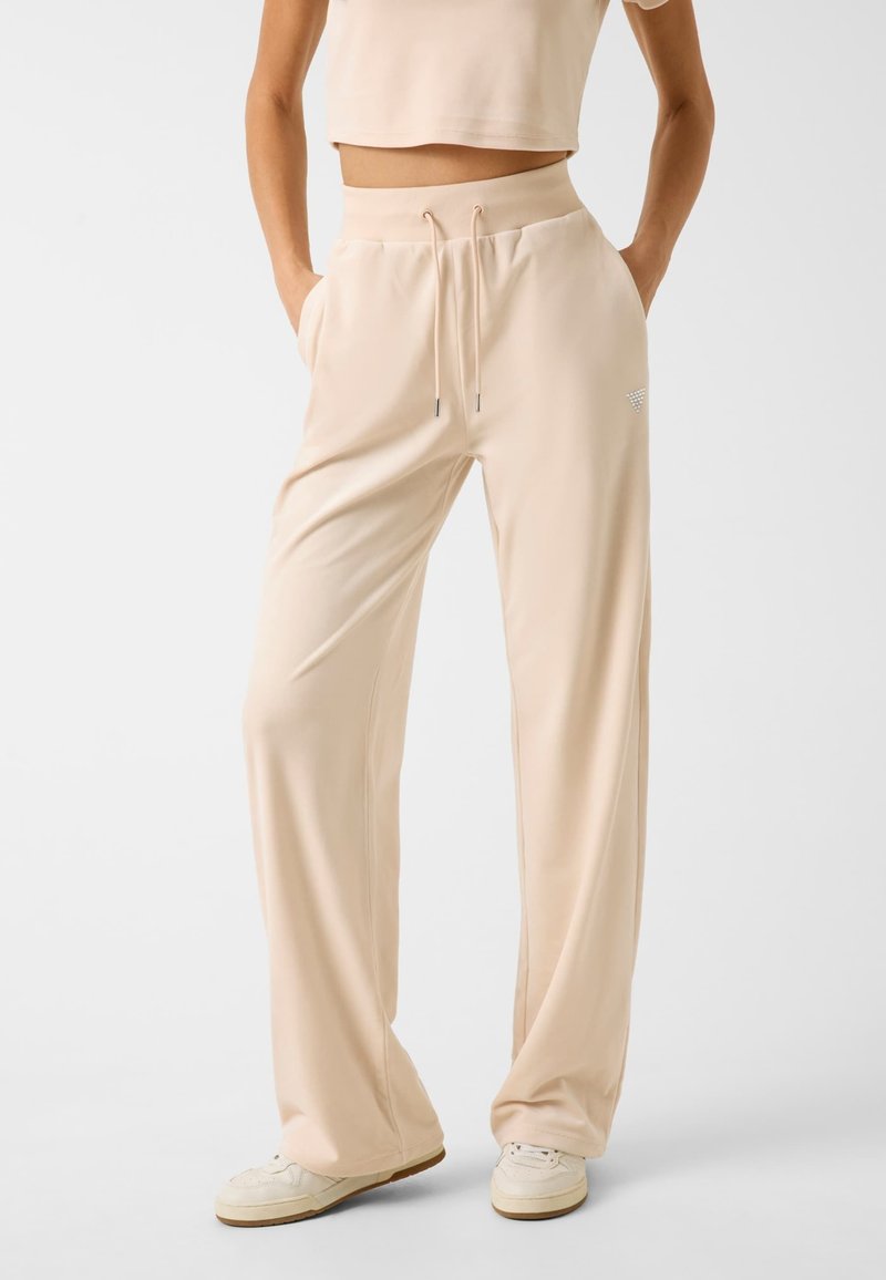 Beige Jersey-Hose mit einem Kordelzugbund, Seitentaschen und einem leichten Glanz, kombiniert mit weißen Sneakers. Elegante und entspannte Passform.
