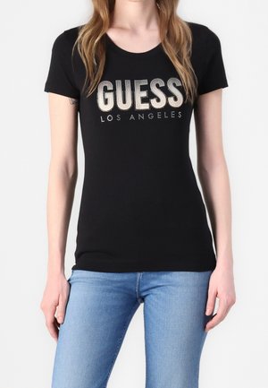 Kobieta ubrana w czarny dopasowany T-shirt z srebrnym napisem "GUESS LOS ANGELES" i jasnoniebieskie jeansy, stojąca na jednolitym białym tle.
