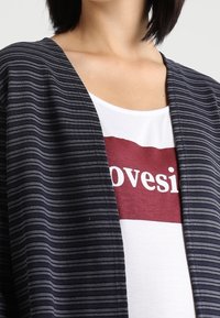Cardigan rayé gris foncé avec un devant ouvert, tissu texturé et coupe décontractée sur un t-shirt blanc avec un graphisme rectangulaire rouge.