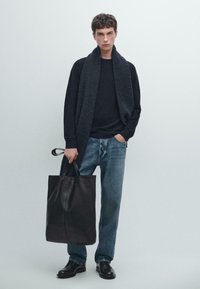 Zwarte leren tote bag, oversized donker sweater, losse lichtblauwe jeans, krullend haar, staand tegen een effen lichte achtergrond.