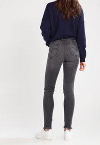 Jeans skinny de color gris oscuro con un dobladillo deshilachado, dos bolsillos traseros y una pequeña etiqueta de marca roja, combinados con un suéter azul marino y zapatillas blancas.
