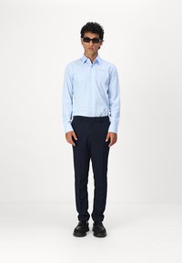 Helles blaues Hemd mit Knöpfen, dunkelblaues Slim-Fit-Hosen, schwarze Schuhe und Sonnenbrille. Das Model steht mit Händen an den Seiten vor einem einfarbigen Hintergrund.