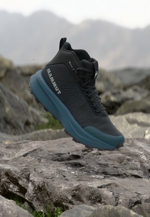 Must sinine Mammut matkasaabas õhus üle kivise maastiku, taustal on mäed uduse fookusega.