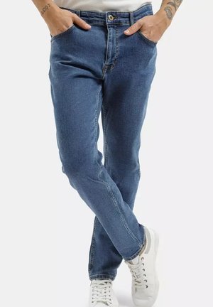 Osoba nosząca średnio niebieskie dżinsy, z rękami w kieszeniach, skrzyżowanymi nogami i białymi trampkami na białym tle.