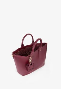 Sac fourre-tout en cuir bordeaux avec quincaillerie dorée, double anses, et une petite pochette attachée. L'intérieur comprend des poches zippées et des poches plaquées.
