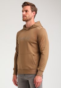 Bruin hoodie met een voorzak, voorzien van een textuurlogo-ontwerp over de borst en geribbelde manchetten, gemaakt van zachte stof.
