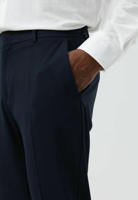 Marineblaue Hosen aus glattem Stoff, mit schmaler Passform, Seitentasche und dezenten Falten, kombiniert mit einem weißen Langarmhemd.