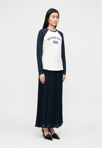 Camisola raglan de mangas compridas branca e azul-marinho com impressão "MICHAEL KORS 1981", combinada com uma saia longa plissada azul-marinho e sapatos pretos de salto.