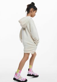 Beige színű kapucnis pulóverruha hosszú ujjal és oldalzsebekkel, rózsaszín szandálokkal és pasztell színű batikolt zoknikkal párosítva. Egyszerű dizájn, laza stílus.