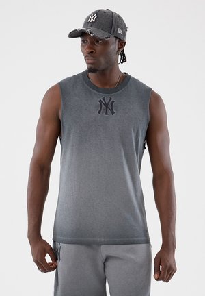 Hombre con una camiseta sin mangas gris, pantalones de chándal a juego y una gorra desgastada de los New York Yankees, mirando hacia su izquierda.