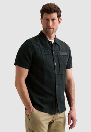 Man with short brown hair wearing short-sleeve black button-up shirt and beige pants, standing with one hand in pocket against plain background.