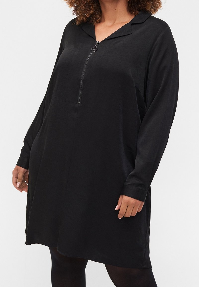 Vestito nero a maniche lunghe con colletto e cerniera frontale. Tessuto morbido, vestibilità ampia, arriva a metà coscia. Presenta tasche laterali e maniche con polsini.