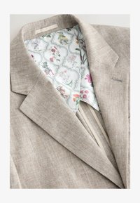 Chaqueta de traje de lino beige con forro de estampado floral. Presenta un exterior texturizado y un corte moderno con solapas de pico.