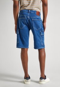 Blå denim cargo shorts med flera fickor, en kontrasterande brun läderlapp bak, och synliga sömdetaljer, bärs med vita sneakers.