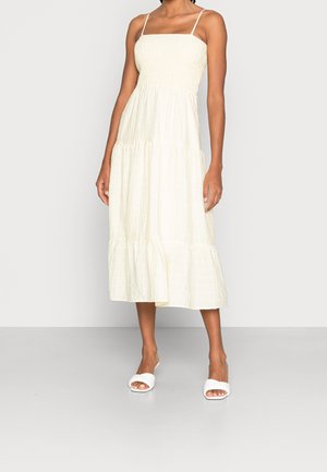 Kobieta ubrana w kremową, midi sukienkę na cienkich ramiączkach z teksturowanej tkaniny, w połączeniu z białymi, otwartymi sandałami na płaskim obcasie.