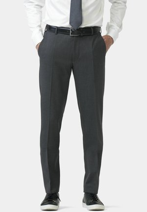 Homme portant un pantalon gris sur mesure avec pli, ceinture noire, chemise blanche, cravate grise, et baskets basses noires à semelles blanches, les mains dans les poches.