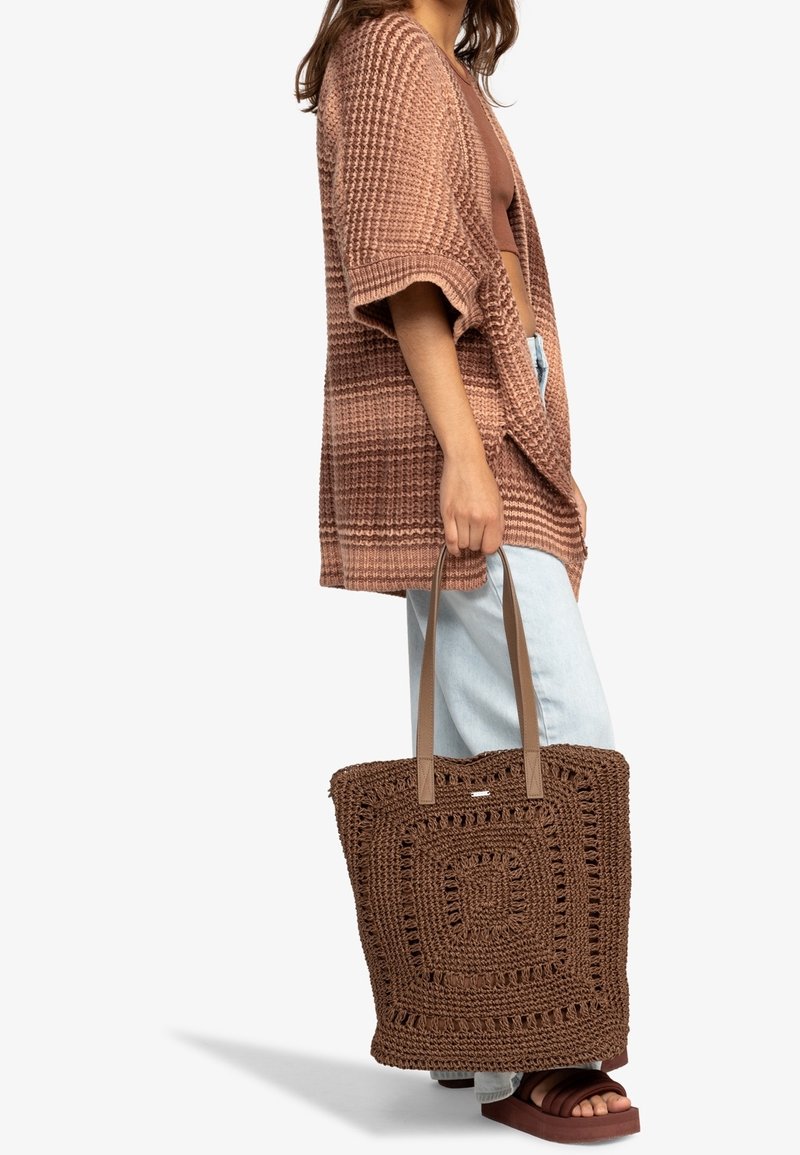 Femme portant un cardigan marron texturé, un jean bleu clair, des sandales marron, tenant un grand sac cabas carré marron en crochet par les poignées.