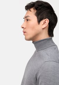 Grauer Rollkragenpullover mit geripptem Kragen, tailliertem Schnitt, glatter Textur und weichem Stoff. Seitenansicht des Kleidungsstücks, das von einem Modell getragen wird.