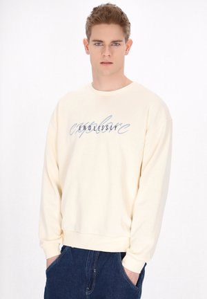 Cremefarvet sweatshirt med lange ærmer, ribbede manchetter og rund halsudskæring. Har "ENDLESS" i blå kursiv tekst på tværs af brystet.