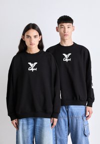Černé oversized mikiny vyrobené z bavlněné směsi, s bílým tištěným logem na hrudi. V kombinaci s modrými džínovými kalhotami.