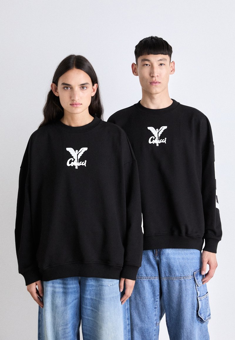 Černé oversized mikiny vyrobené z bavlněné směsi, s bílým tištěným logem na hrudi. V kombinaci s modrými džínovými kalhotami.