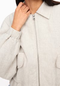 Light gray linen jacket with a zip front, open collar, and gathered sleeves. Features two front pockets and textured fabric.
